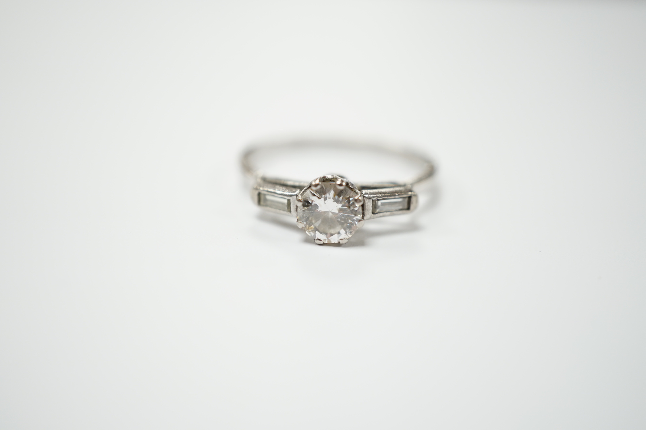 A white metal and single stone diamond set ring, with two stone baguette cut diamond set shoulders, size O, gross weight 2.3 grams, stone diameter 5.2mm.